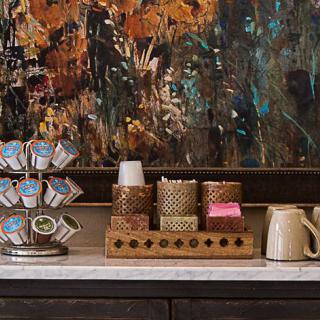 A cozy coffee station with assorted pods, mugs, and rustic decor against a colorful abstract painting.