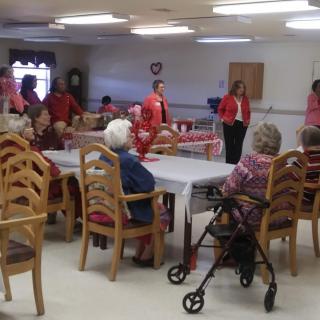 A group of elderly people gathered in a community room, some sitting around tables and others standing in conversation.