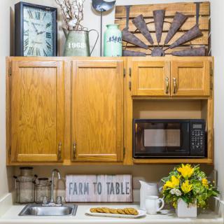 A charming farmhouse-style kitchen setup with wooden cabinets, a microwave, and decorative elements including a clock, flowers, and a 'Farm to Table' sign.