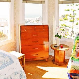 A cozy bedroom with natural light streaming through large windows, featuring a wooden dresser and round side table.