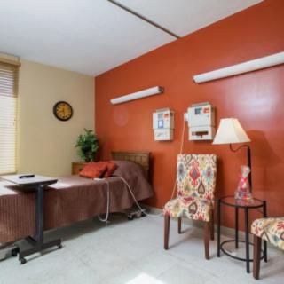 A comfortable nursing home or hospital room featuring a bed, two chairs, a side table with a lamp, and warm-colored walls.