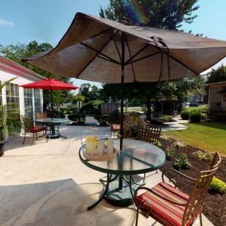 A sunny patio with a glass table, chairs, and large umbrellas surrounded by greenery.