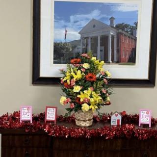 A decorative floral arrangement placed on a table, surrounded by festive red garland and small gifts, with a painting of a building in the background.
