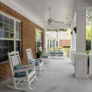 A cozy porch featuring white rocking chairs with cushions, a ceiling fan, and brick walls.