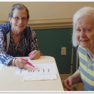 Two elderly women smiling while sitting at a table with papers.