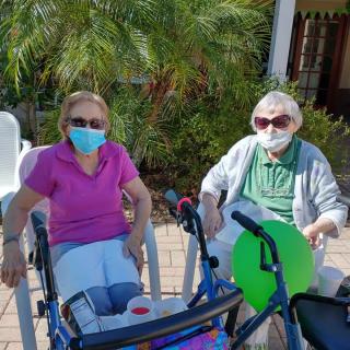 Two individuals sitting outdoors in wheelchairs, wearing masks and sunglasses, surrounded by plants.