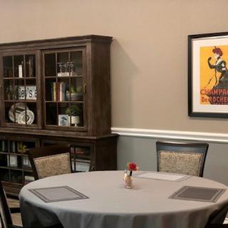 A cozy dining room with a round table, cushioned chairs, a wooden bookshelf, and wall artwork.