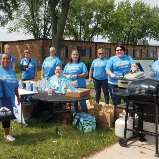 A group of people gathered outdoors around a table and a barbecue grill, participating in a community event.