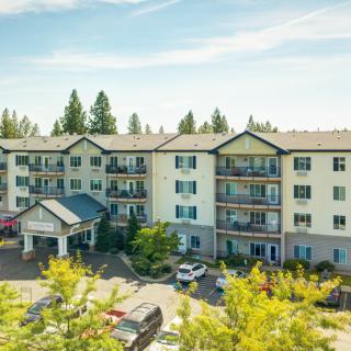 A multi-story apartment building with a parking lot surrounded by trees under a blue sky.