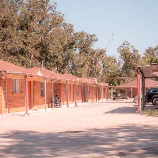 A row of motel rooms with a parking area and bicycles, surrounded by trees during the day.