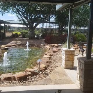 A serene outdoor space featuring a pond with fountains, surrounded by lush greenery and stone landscaping.