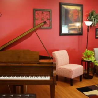A cozy living room with a grand piano against a red wall, decorated with artwork, chairs, and a floor lamp.