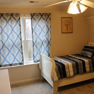 A cozy bedroom with a single bed featuring striped bedding, curtains on the window, and a ceiling fan.