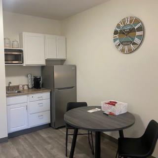 A kitchenette with a small dining area, featuring a table and chairs, fridge, microwave, cabinets, and a decorative wall clock.