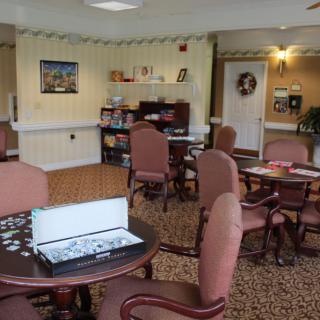 A cozy recreation room with tables, chairs, a puzzle in progress, and a bookshelf.