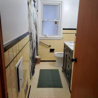 A narrow bathroom with yellow tile walls, a window, and a green rug on the tile floor.