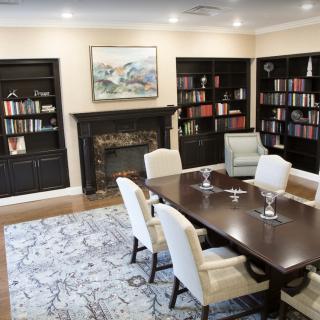 A cozy library room featuring a large wooden table and chairs, surrounded by bookshelves filled with books. A fireplace and artwork adorn the wall.