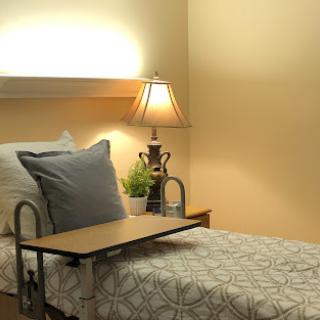 A cozy hospital room featuring a neatly made bed, a bedside lamp, and a small table tray.