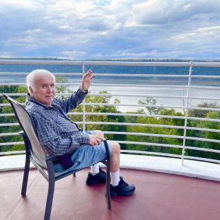 An elderly man sits on a balcony, enjoying a scenic ocean view under a cloudy sky.