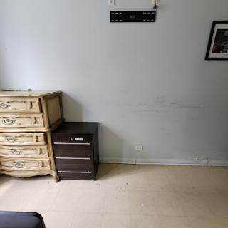 A minimalist room featuring a light-colored wall, a set of drawers, and a dark nightstand.
