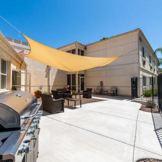 A sunny residential backyard with a patio, featuring a barbecue grill and outdoor furniture under a shade sail.