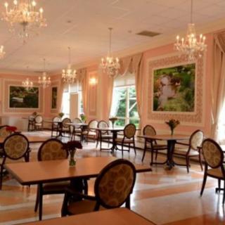 An elegant dining area adorned with chandeliers, featuring neatly arranged tables and chairs, large windows letting in natural light, and classic paintings on the walls.