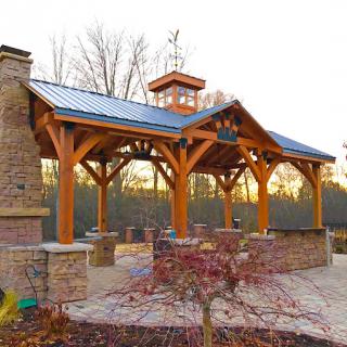 An outdoor timber frame pergola with a built-in fireplace, set against a natural landscape.