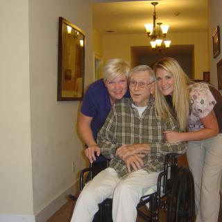 An elderly person in a wheelchair smiles with two caregivers by their side in a hallway.