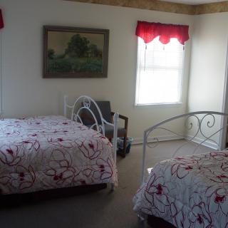 A cozy bedroom featuring two metal frame beds with floral bedding, a window with red valance, and a landscape painting on the wall.