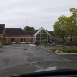 A view of a building with a parking lot in front, surrounded by trees under a cloudy sky.