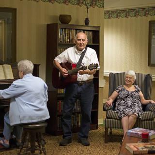 A man plays the guitar while a woman plays the piano and another woman sits in a chair, enjoying the music in a cozy living room setting.