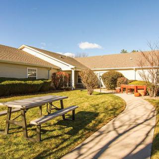 A sunny outdoor area with a pathway leading to a house, surrounded by a garden, benches, and picnic tables.