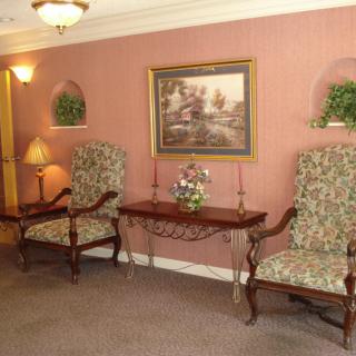 A traditionally styled interior with floral upholstered chairs, a wooden table, decorative plants, and a painting on the wall.