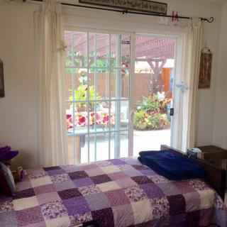 A cozy bedroom with a purple quilt on the bed, overlooking a garden through a sliding glass door.