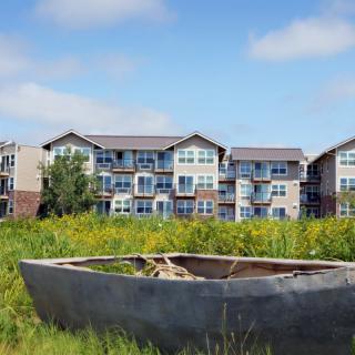 An abandoned boat rests in a grassy field with an apartment building in the background against a blue sky.