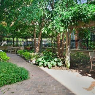 A peaceful garden pathway surrounded by lush greenery and shaded by trees, featuring a bench and a fence.