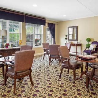 A cozy room with several tables and chairs, featuring a patterned carpet and large windows with curtains.