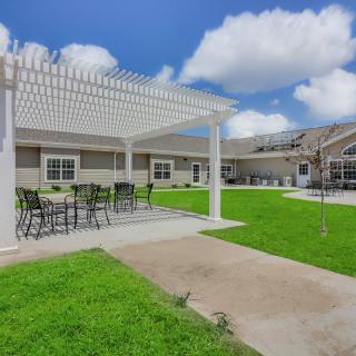 A spacious outdoor patio area with a pergola and seating, surrounded by green lawn and under a clear blue sky with scattered clouds.