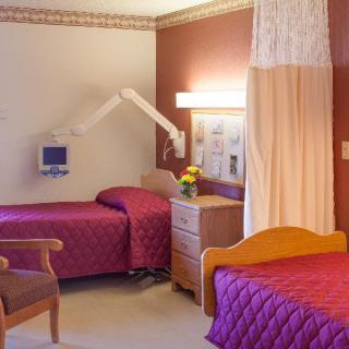 A hospital room featuring two beds with quilted covers, a chair, medical equipment on the wall, and a small table with flowers and a lamp.