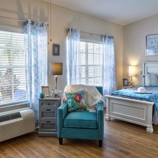 A cozy and well-lit bedroom featuring a blue armchair, elegant curtains, and wooden flooring.