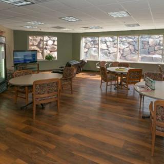 A spacious lounge area with wooden flooring, featuring several tables and chairs, large windows, and a television.