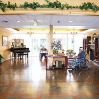 A cozy interior of a nursing home with residents seated, featuring a piano and decorative plants.