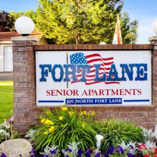 A brick sign for Fort Lane Senior Apartments featuring an American flag, surrounded by landscaping and trees.