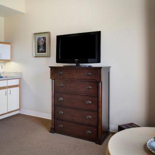 A room with a chest of drawers topped by a television, adjacent to a kitchenette.