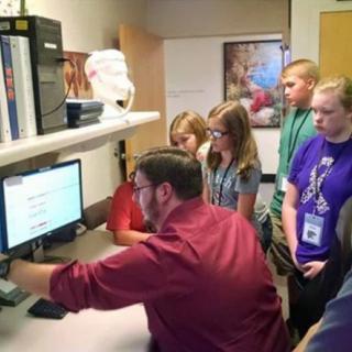 A group of students gathers around a computer as an instructor demonstrates something on the screen.