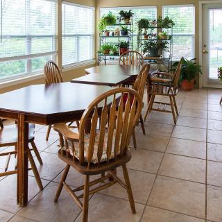 A bright dining area with wooden tables and chairs, surrounded by indoor plants and large windows letting in natural light.