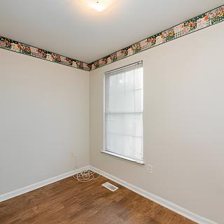 A small, empty room with a hardwood floor and a single window, featuring a wallpaper border near the ceiling.