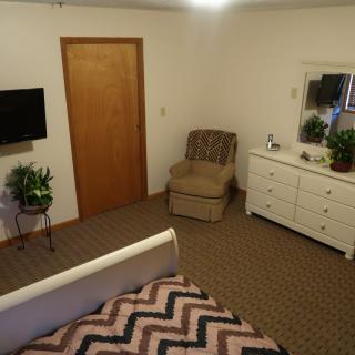 A cozy bedroom interior with a bed, armchair, dresser, and television.