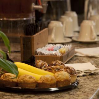 A selection of pastries and bananas on a countertop, accompanied by coffee cups and sugar packets.