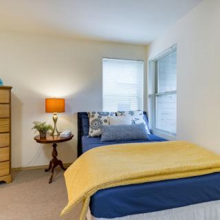 A cozy bedroom with a neatly made bed, a side table with a lamp, and a dresser by the window.